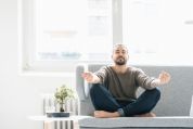 Mann mit rundem Gesicht, dünnem Vollbart, geschlossenen Augen und blossen Füssen sitzt im Schneidersitz auf einem Sofa, die Arme ausgestreckt - neben sich auf einem Tischchen ein Bonsai - und tut so, als wenn er meditiert Mann mit rundem Gesicht, dünnem Vollbart, geschlossenen Augen und blossen Füssen sitzt im Schneidersitz auf einem Sofa, die Arme ausgestreckt - neben sich auf einem Tischchen ein Bonsai - und tut so, als wenn er meditiert